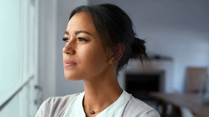 Close-up of a woman in a gray sweater, hair pulled back, looking out the window with a thoughtful expression. The blurred modern living room suggests her focus on handling probate real estate mistakes. Contact Kid Breukelen Realty Group for guidance.