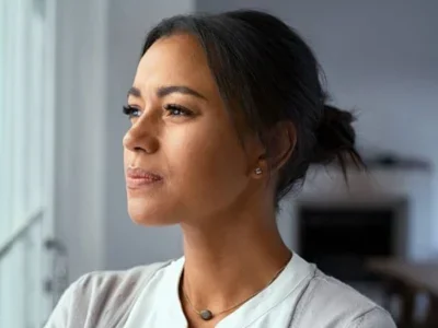 Woman in a gray sweater with hair pulled back, looking out the window in a blurred modern living room, reflecting on probate real estate mistakes. Contact Kid Breukelen Realty Group for expert assistance.