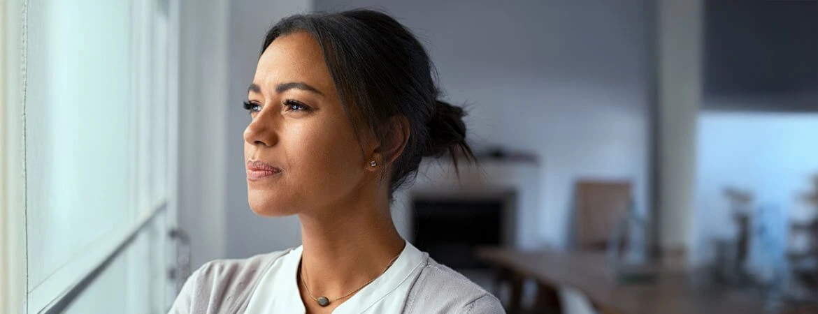 A close-up of a woman in a gray sweater, her hair pulled back, gazing thoughtfully out the window. Behind her, a blurred modern living room suggests her focus on making informed decisions regarding probate real estate mistakes. Reach out to Kid Breukelen Realty Group for expert help.