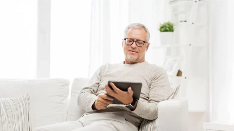 Man sitting on a white sofa, reading a tablet in a white-themed room, reviewing essential probate real estate terms. Contact Kid Breukelen Realty Group for assistance.